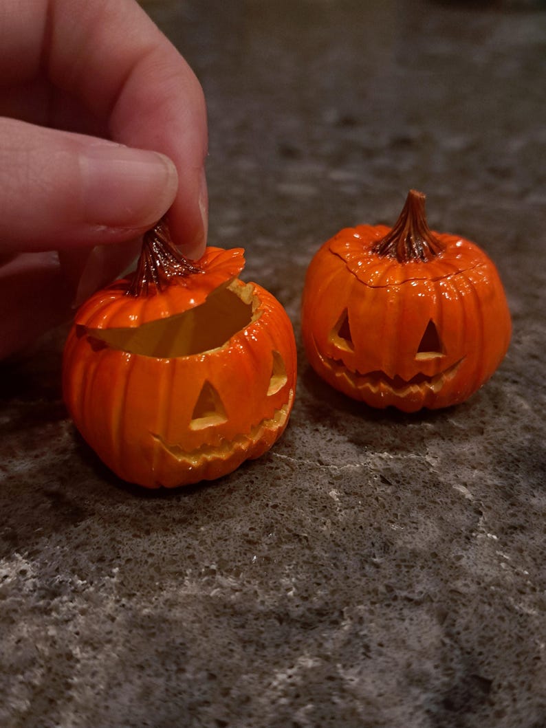 May include: Two small orange jack-o'-lanterns with carved triangular eyes and jagged mouths. One pumpkin is shown with its lid slightly lifted, revealing a hollow interior, while the other pumpkin remains closed with a dark brown stem on top.