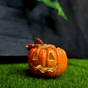 May include: A bright orange ceramic jack-o'-lantern figurine with a glossy finish. The pumpkin features carved triangular eyes and a jagged, toothy grin, topped with a textured brown stem. The object is hollow and has a lid-like top section.