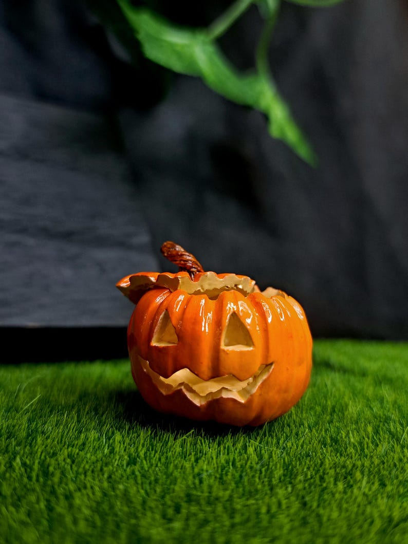 May include: A bright orange ceramic jack-o'-lantern figurine with a glossy finish. The pumpkin features carved triangular eyes and a jagged, toothy grin, topped with a textured brown stem. The object is hollow and has a lid-like top section.