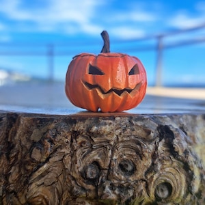 May include: A small, orange, carved pumpkin figurine with a dark brown stem. The pumpkin features a carved, jagged, grinning face with triangular eyes and a wide, toothy mouth, creating a classic jack-o'-lantern appearance with a smooth finish.