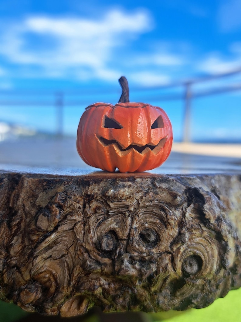 May include: A small, orange, carved pumpkin figurine with a dark brown stem. The pumpkin features a carved, jagged, grinning face with triangular eyes and a wide, toothy mouth, creating a classic jack-o'-lantern appearance with a smooth finish.