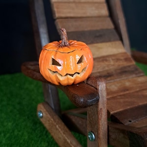 May include: A small, bright orange ceramic pumpkin with a carved jack-o'-lantern face featuring triangular eyes and a jagged mouth. The pumpkin has a textured brown stem and sits on the armrest of a miniature wooden lounge chair.
