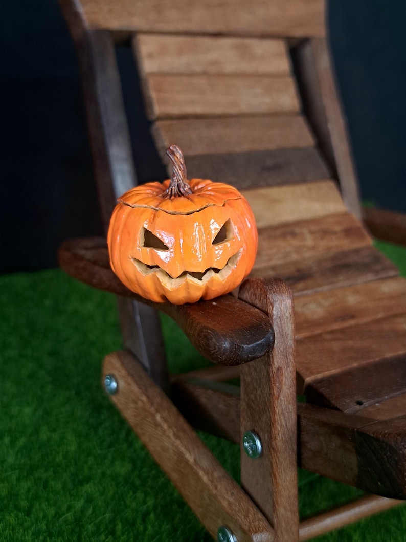 May include: A small, bright orange ceramic pumpkin with a carved jack-o'-lantern face featuring triangular eyes and a jagged mouth. The pumpkin has a textured brown stem and sits on the armrest of a miniature wooden lounge chair.