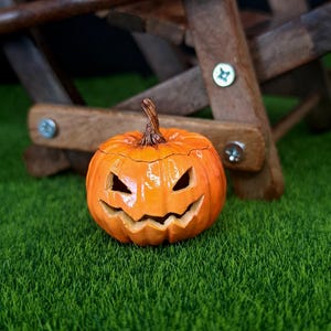 May include: A bright orange ceramic jack-o'-lantern with a glossy finish, featuring carved triangular eyes and a jagged, grinning mouth. The pumpkin has a textured brown stem at the top and a rounded, ribbed body with deep vertical grooves.