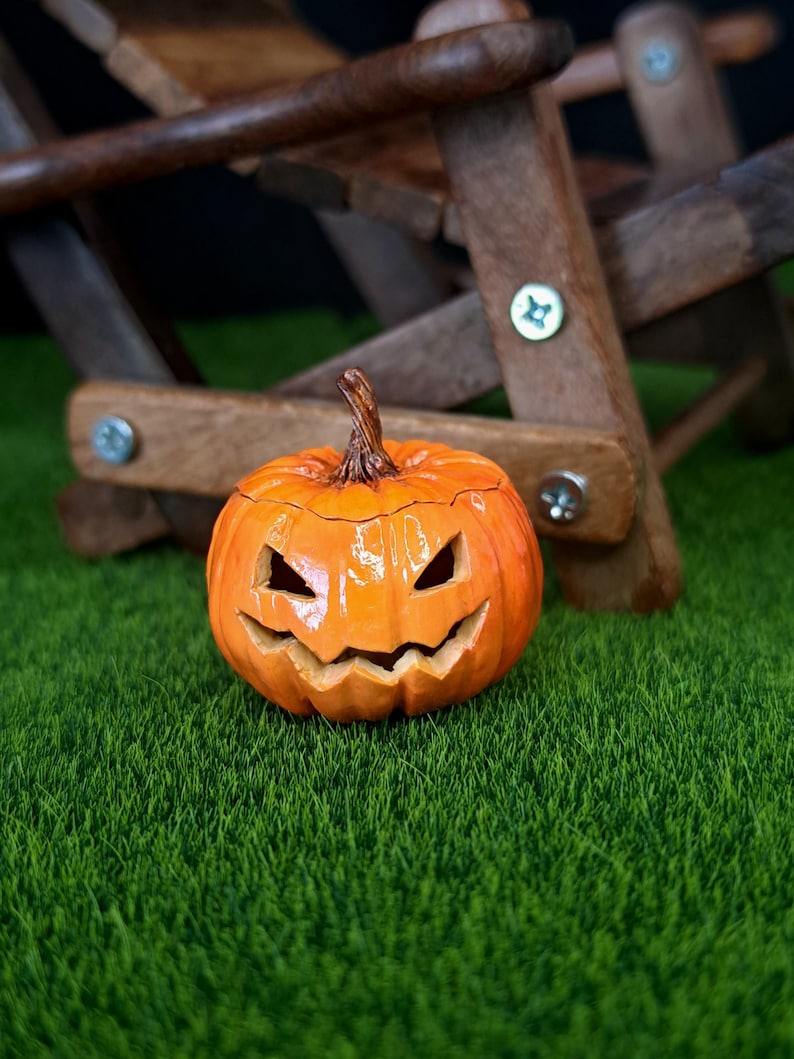 May include: A bright orange ceramic jack-o'-lantern with a glossy finish, featuring carved triangular eyes and a jagged, grinning mouth. The pumpkin has a textured brown stem at the top and a rounded, ribbed body with deep vertical grooves.