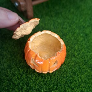 May include: A miniature orange jack-o'-lantern container with a removable lid. The pumpkin-shaped vessel features carved facial cutouts and a glossy finish, with the lid held separately to reveal the hollow interior of the small decorative item.