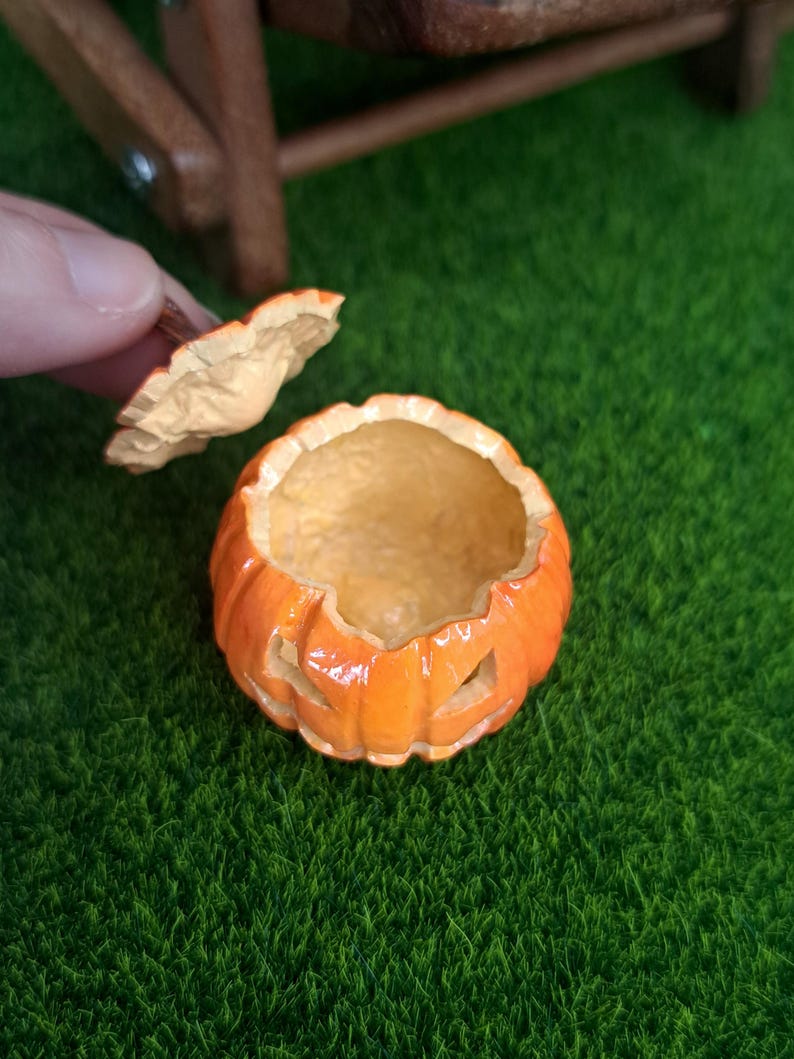 May include: A miniature orange jack-o'-lantern container with a removable lid. The pumpkin-shaped vessel features carved facial cutouts and a glossy finish, with the lid held separately to reveal the hollow interior of the small decorative item.
