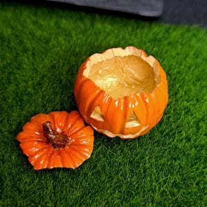 May include: A small, bright orange jack-o'-lantern container with a removable lid. The pumpkin-shaped vessel features carved triangular eyes and a mouth, with a glossy finish and a textured brown stem on the lid, revealing a hollow interior.