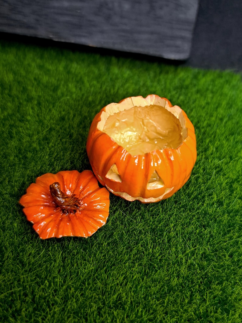 May include: A small, bright orange jack-o'-lantern container with a removable lid. The pumpkin-shaped vessel features carved triangular eyes and a mouth, with a glossy finish and a textured brown stem on the lid, revealing a hollow interior.