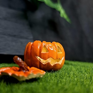 May include: A glossy orange ceramic jack-o'-lantern container with a removable lid. The pumpkin features carved triangular eyes and a jagged mouth, with a brown stem on the lid. The surface is smooth and reflective, showing a bright orange finish.