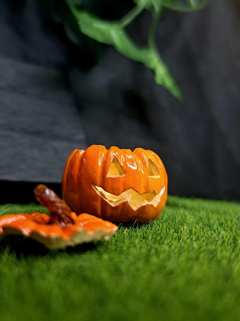 May include: A glossy orange ceramic jack-o'-lantern container with a removable lid. The pumpkin features carved triangular eyes and a jagged mouth, with a brown stem on the lid. The surface is smooth and reflective, showing a bright orange finish.