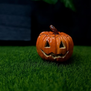 May include: A small, glossy orange jack-o'-lantern figurine with a carved face featuring triangular eyes and a jagged, smiling mouth. The pumpkin has a textured brown stem at the top and a rounded, ribbed body with deep vertical grooves.