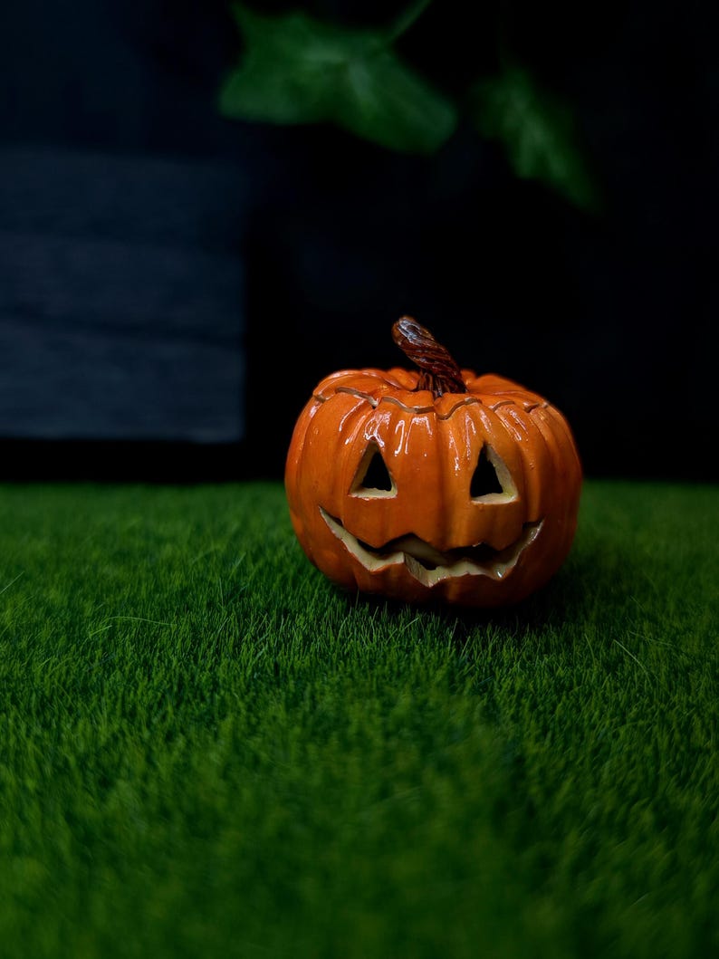 May include: A small, glossy orange jack-o'-lantern figurine with a carved face featuring triangular eyes and a jagged, smiling mouth. The pumpkin has a textured brown stem at the top and a rounded, ribbed body with deep vertical grooves.