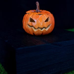 May include: A carved orange pumpkin jack-o'-lantern with a glossy finish, featuring triangular eyes and a jagged, toothy grin. The pumpkin has a textured brown stem at the top and deep vertical ridges along its round, bulbous body.