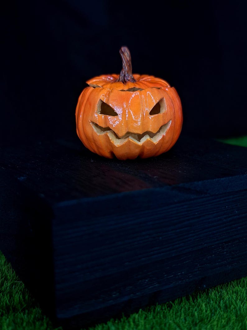 May include: A carved orange pumpkin jack-o'-lantern with a glossy finish, featuring triangular eyes and a jagged, toothy grin. The pumpkin has a textured brown stem at the top and deep vertical ridges along its round, bulbous body.