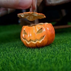 May include: A small orange jack-o'-lantern container with a removable lid. The pumpkin features a carved, grinning face with triangular eyes and a jagged mouth. The surface has a glossy finish, and the lid is held slightly above the base.