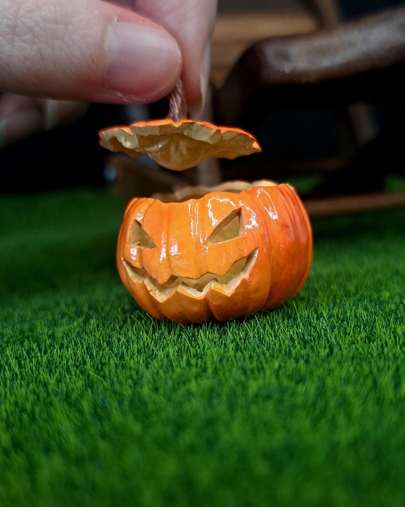 May include: A small orange jack-o'-lantern container with a removable lid. The pumpkin features a carved, grinning face with triangular eyes and a jagged mouth. The surface has a glossy finish, and the lid is held slightly above the base.