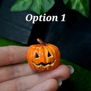 May include: A miniature orange jack-o'-lantern pumpkin figurine with a carved triangular eye and jagged mouth expression, featuring a textured brown stem on top. The small decorative object displays a glossy finish and vertical ridges.