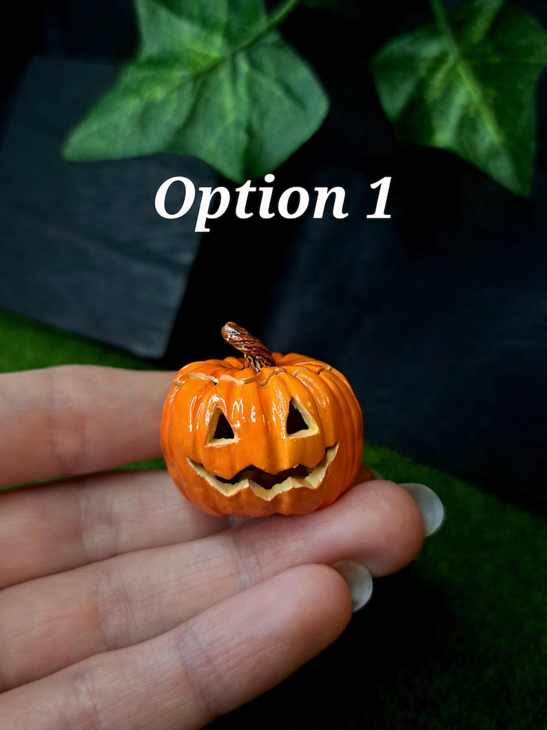 May include: A miniature orange jack-o'-lantern pumpkin figurine with a carved triangular eye and jagged mouth expression, featuring a textured brown stem on top. The small decorative object displays a glossy finish and vertical ridges.