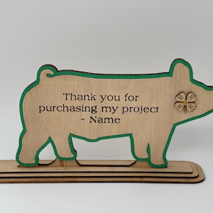 May include: Wooden pig cutout with a green outline and a 4-H clover on the side. The pig is standing on a wooden base. The cutout has the text "Thank you for purchasing my project ~ Name" on it.