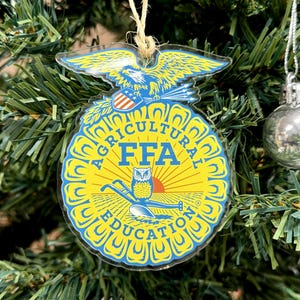 May include: A clear FFA ornament with a yellow and blue design, hanging on a Christmas tree. The ornament features an eagle, the FFA logo, and the words "AGRICULTURAL EDUCATION". A silver ball ornament is also visible.