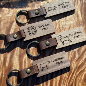 May include: Three wooden keychains with brown leather straps. Each keychain features a different animal silhouette: a pig, a goat, and a cow. The keychains are engraved with the text "Custom Text". One keychain also features a 4-H clover logo.