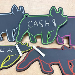 May include: A collection of chalkboard pig-shaped signs with colorful outlines. One sign says "CASH" with a dollar sign, and another says "LUCK". Each sign has a hole for hanging and comes with a piece of chalk.