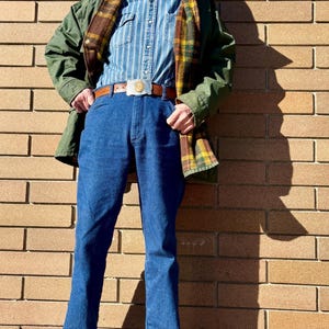 May include: A person wearing a dark beanie, glasses, and a mustache, with a green jacket, plaid scarf, blue striped shirt, blue jeans, brown belt with a silver buckle, and brown shoes. The person is standing in front of a brick wall.