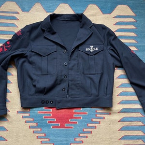 Peut inclure: Veste bleu marine avec fermeture boutonnée, deux poches de poitrine et écussons sur les manches. La veste a un col et le texte "RN.X.S." est visible sur la poitrine. Détails brodés rouges et blancs sur les manches.