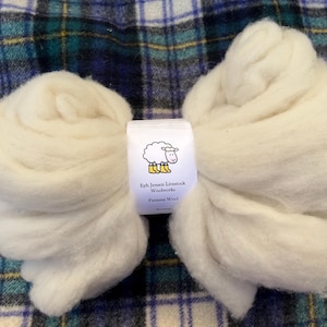 May include: Two large, cream-colored bundles of wool are displayed on a plaid fabric. A label with a sheep illustration and text reading "Eph Jensen Livestock Woolworks Panama Wool" is centered between the bundles. The wool appears soft and fluffy.