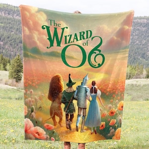 May include: A large, rectangular blanket featuring the iconic imagery from "The Wizard of Oz." The design showcases the characters walking down a yellow brick road, with the title text in green. The background is a vibrant sunset over a field of poppies.