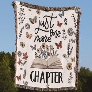 May include: A white woven throw blanket with a book, flowers, and butterflies printed on it. The text "just one more chapter" is printed on the blanket.