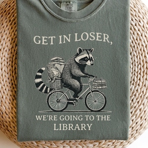 May include: Sage green t-shirt with the text "GET IN LOSER, WE'RE GOING TO THE LIBRARY." A raccoon illustration rides a bicycle carrying books. The shirt is folded on a woven, round surface.