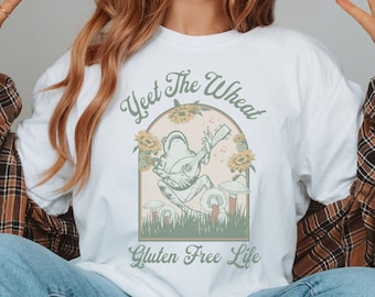 May include: White t-shirt with a green frog playing a banjo in a field of sunflowers. The text "Yeet the Wheat Gluten Free Life" is printed on the shirt.