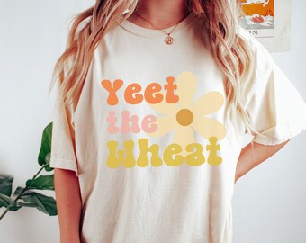May include: A white oversized t-shirt with the text "Yeet the Wheat" in a retro font with a yellow daisy graphic.