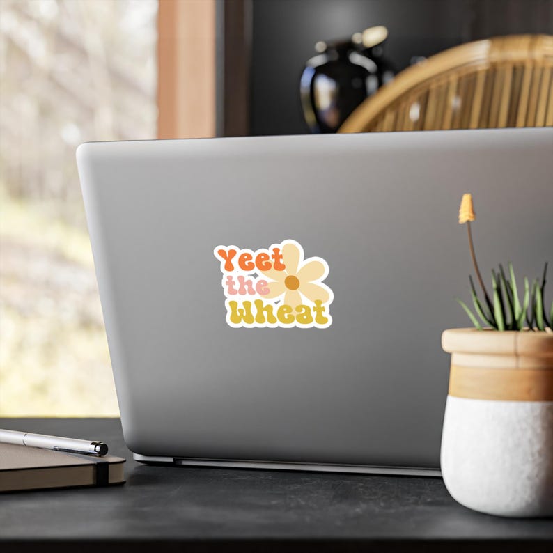May include: A laptop sticker featuring a yellow daisy and the text "Yeet the wheat" in a retro font with orange, pink, and yellow colours.