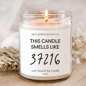 May include: A lit, clear glass jar candle with a white label. The label reads "THIS CANDLE SMELLS LIKE 37216" and "100% Natural Soy Candle 9 oz." The candle has a wooden wick and is on a wooden surface.