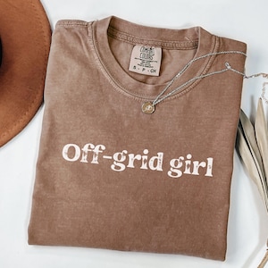 May include: A brown Comfort Colors t-shirt with the text "Off-grid girl" in white. A silver necklace with a pendant is draped across the shirt. The shirt is size small.