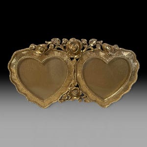 May include: A double heart-shaped picture frame in a metallic gold finish. The frame features a textured border and is decorated with sculpted rose motifs at the top and center. The two heart-shaped openings are set side by side.