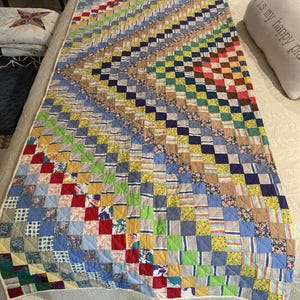 May include: A rectangular patchwork quilt featuring a grid of small, multi-colored fabric squares in various patterns, including floral, striped, and solid prints. The quilt displays a diagonal color gradient pattern across its surface.