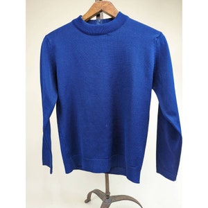 Puede incluir: Un suéter azul real de manga larga con cuello alto. El suéter está hecho de una tela de punto fino y está colgado de una percha de madera. La prenda tiene un diseño clásico y sencillo.