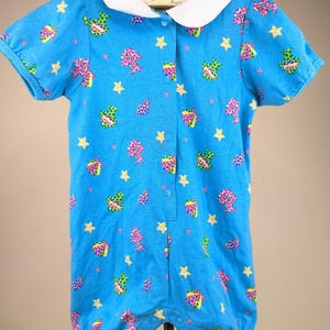 May include: A baby's romper in a vibrant blue hue, adorned with a playful pattern of colorful shapes and yellow stars. The romper features a white Peter Pan collar and short sleeves. The garment is buttoned down the front.