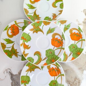 May include: Five white saucers with a floral design. The pattern features orange and green flowers with gold-coloured stems and leaves. The saucers are arranged in a circular pattern on a white marble surface.