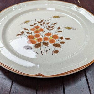 Pode incluir: Um prato de jantar de cerâmica branca com uma borda marrom e um design floral com flores laranja e marrom e espigas de trigo.