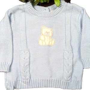May include: A light blue knitted sweater with a cable knit design and a white embroidered teddy bear on the front.