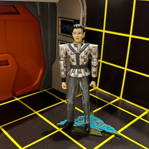 Könnte beinhalten: Eine schwarz-graue Actionfigur einer Person, die ein kariertes Hemd und eine schwarze Hose trägt. Die Figur steht auf einem blauen und grünen Sockel.