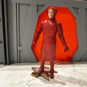 May include: A red action figure of a person wearing a long, hooded robe stands on a gold star-shaped base. The figure has articulated arms and legs.