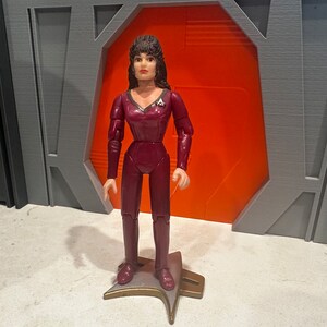 May include: A maroon action figure of a woman in a Star Trek uniform, standing on a gold and silver base. The figure has dark hair and is posed in front of a gray and orange backdrop. The figure is approximately 6 inches tall.