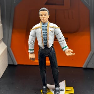 May include: A white and gold Star Trek action figure of a character wearing a white uniform with gold accents. The figure is standing on a black and gold base.