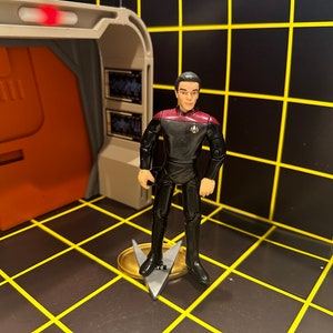Op de afbeelding: Actiefiguur van een personage in een zwart uniform met een bordeauxrode kraag, staand op een goud-zilveren basis. De figuur bevindt zich in een futuristische omgeving met oranje en grijze muren en een zwarte vloer met gele rasterlijnen.
